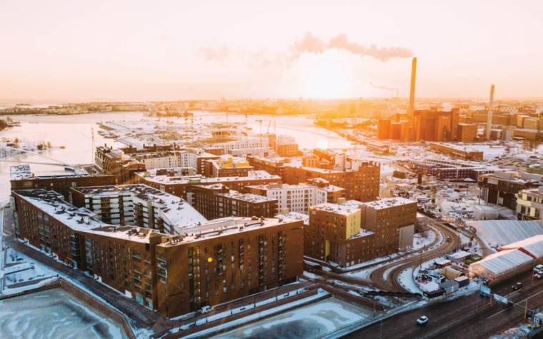 An aerial photo of Helsinki, Finland. Buildings, road, smoke coming from a chimney of a power plant, snow on the ground. A shining sun.