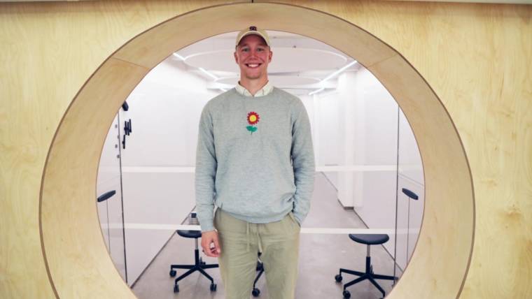 A young man standing in fron of a circular window indoors. The young man wears a grey sweater with a flower print, beige cap and trousers. Behind the window an aisle with two stools. 